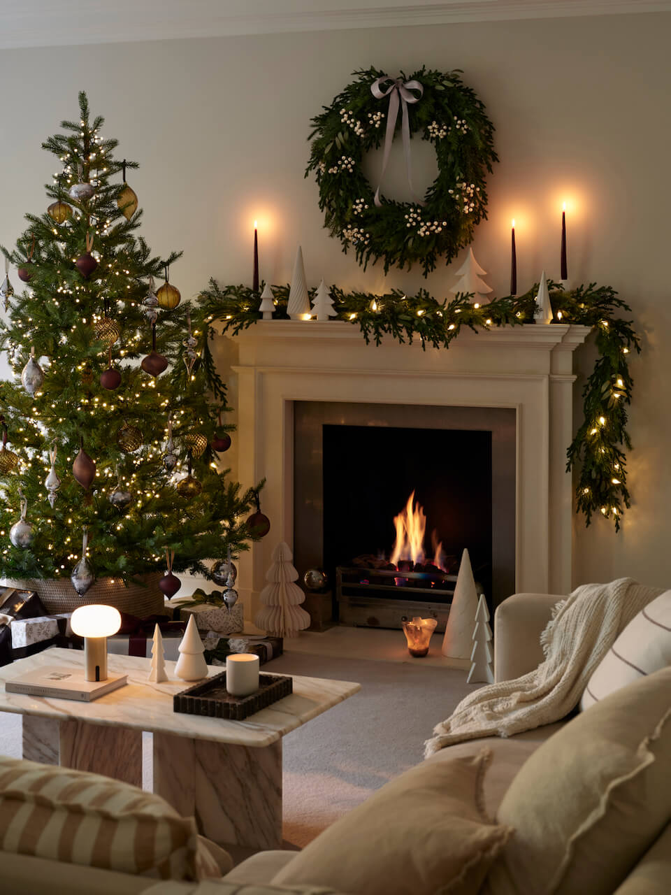 Christmas living room with a decorated tree, foliage wreath, mantelpiece garland and white ceramic Christmas trees.