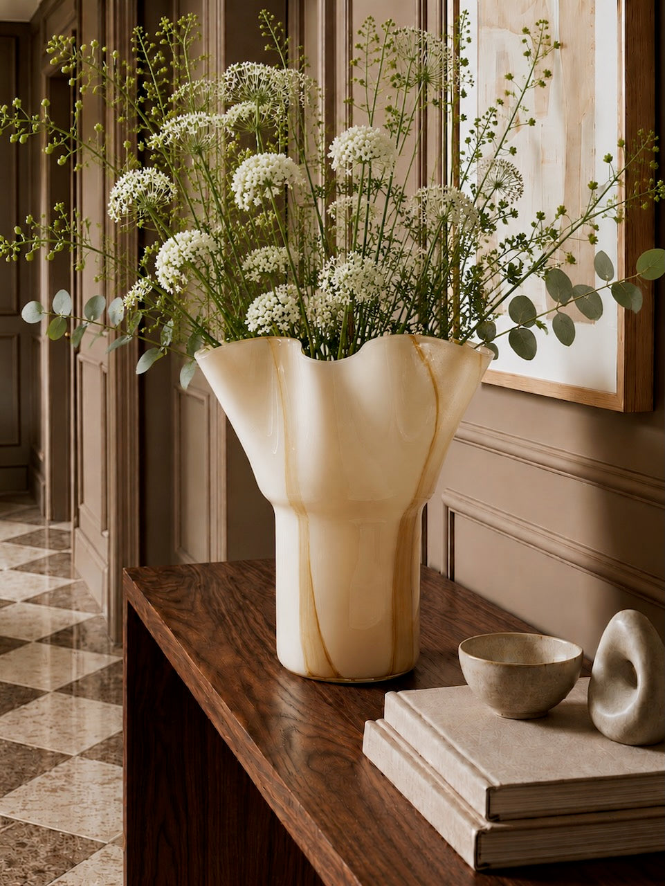 Decorative vase with flowers on a wooden shelf in a room with a checkered floor.