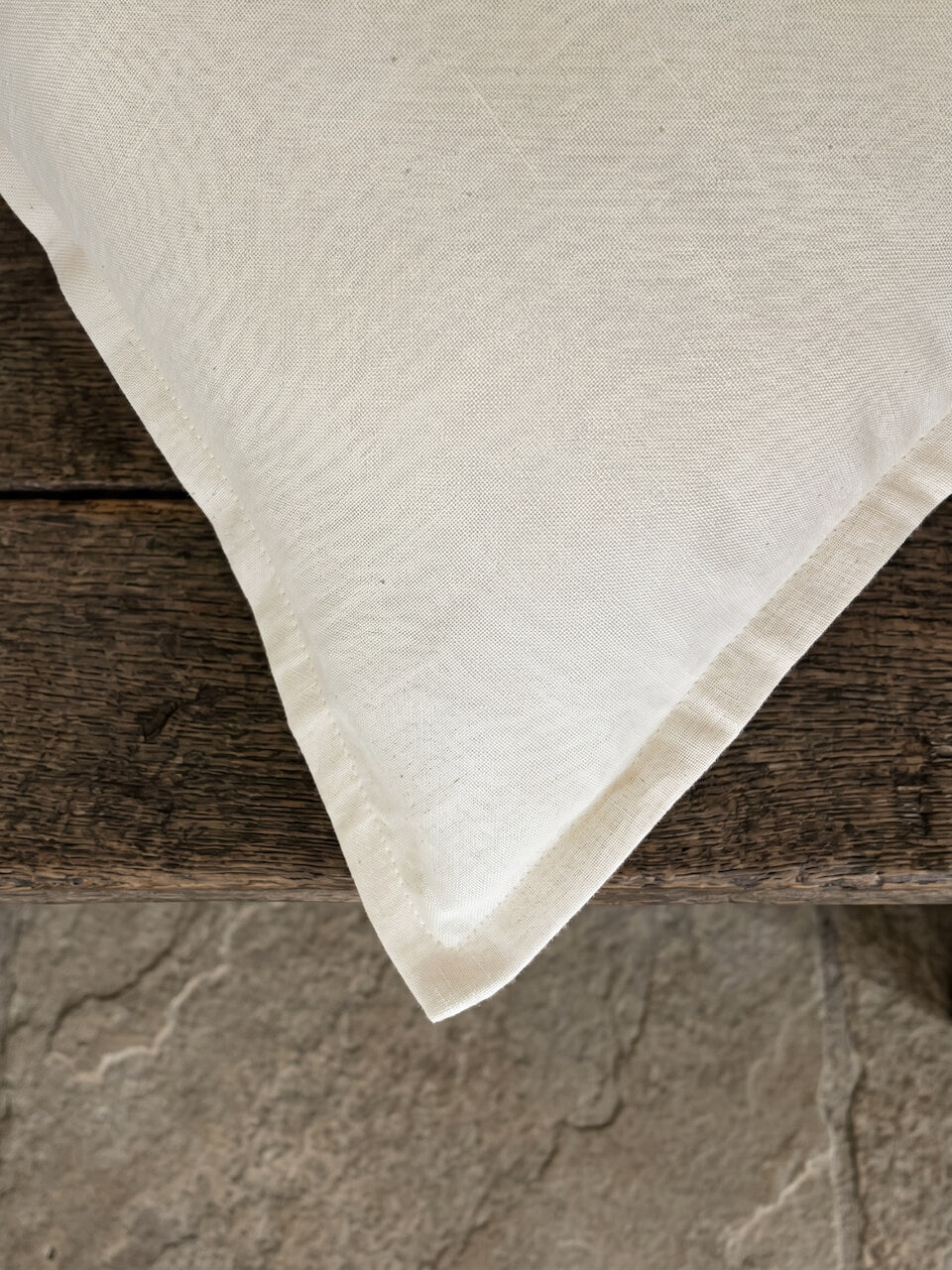 Close-up detail of a luxury cream cotton linen cushion cover featuring a soft woven texture and tailored edging, styled on rustic timber for a refined contemporary aesthetic.