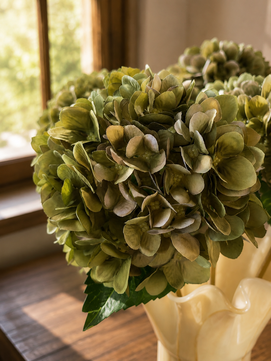 Close-up detail of vintage green faux hydrangea blooms showcasing realistic layered petals and natural colouring, arranged in a cream marbled ruffle vase with soft spring sunlight filtering through a wooden window.