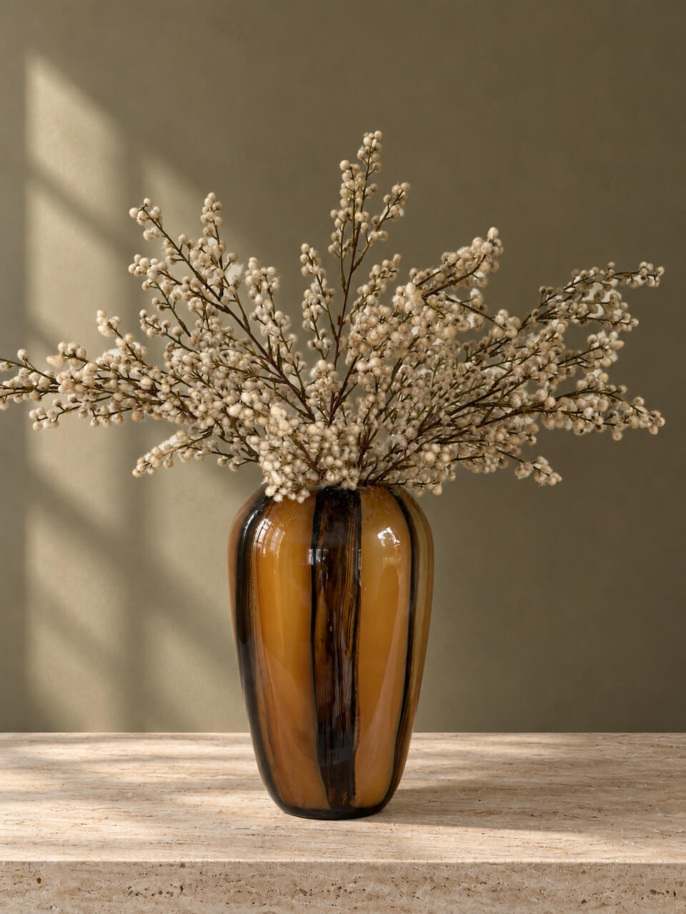 A glossy amber-brown glass vase with dark vertical streaks, filled with dried white baby's breath flowers, set on a light stone surface against a warm taupe wall with soft window shadows.