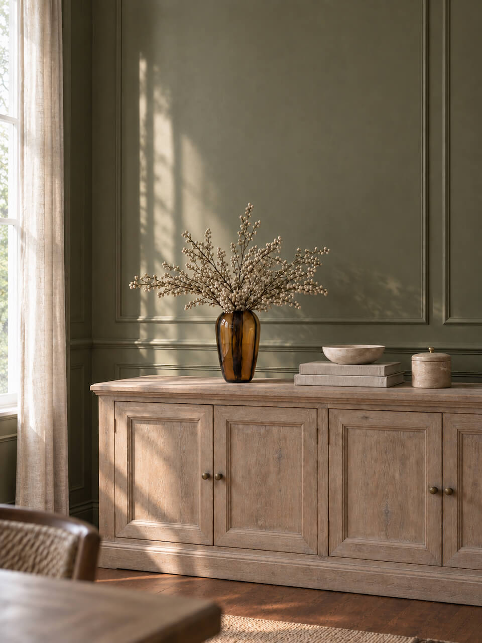 A rustic light wood sideboard or credenza in a styled room, decorated with a tall amber glass vase holding dried white branches, a small bowl, and a jar. The room features a sage green paneled wall and warm natural light from a window.