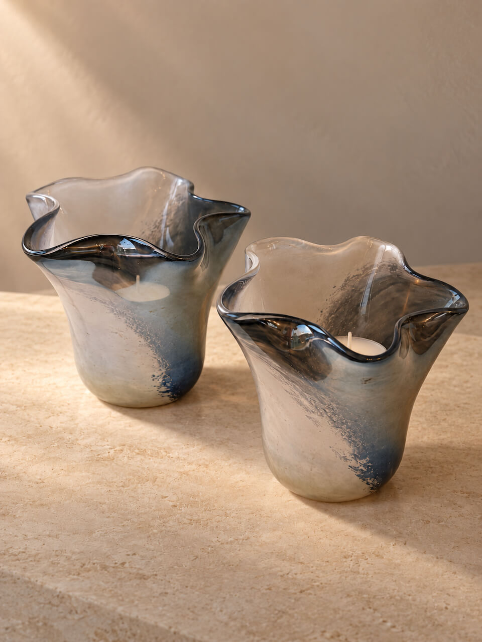 Two sculptural, wavy-edge glass candle holders with a smoky gray and navy blue marbled finish, displayed on a textured beige stone surface against a neutral beige background.