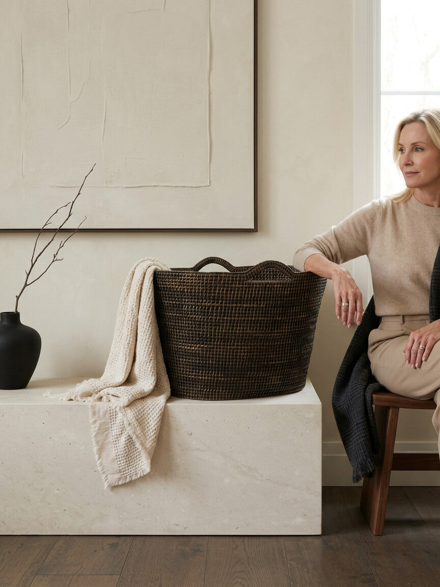 Nairn extra large brown rattan bucket basket styled with blanket on stone bench in warm neutral living space. The basket has a throw flowing out o fit on one side and on the other side a blond lady has her elbow leaning on it.