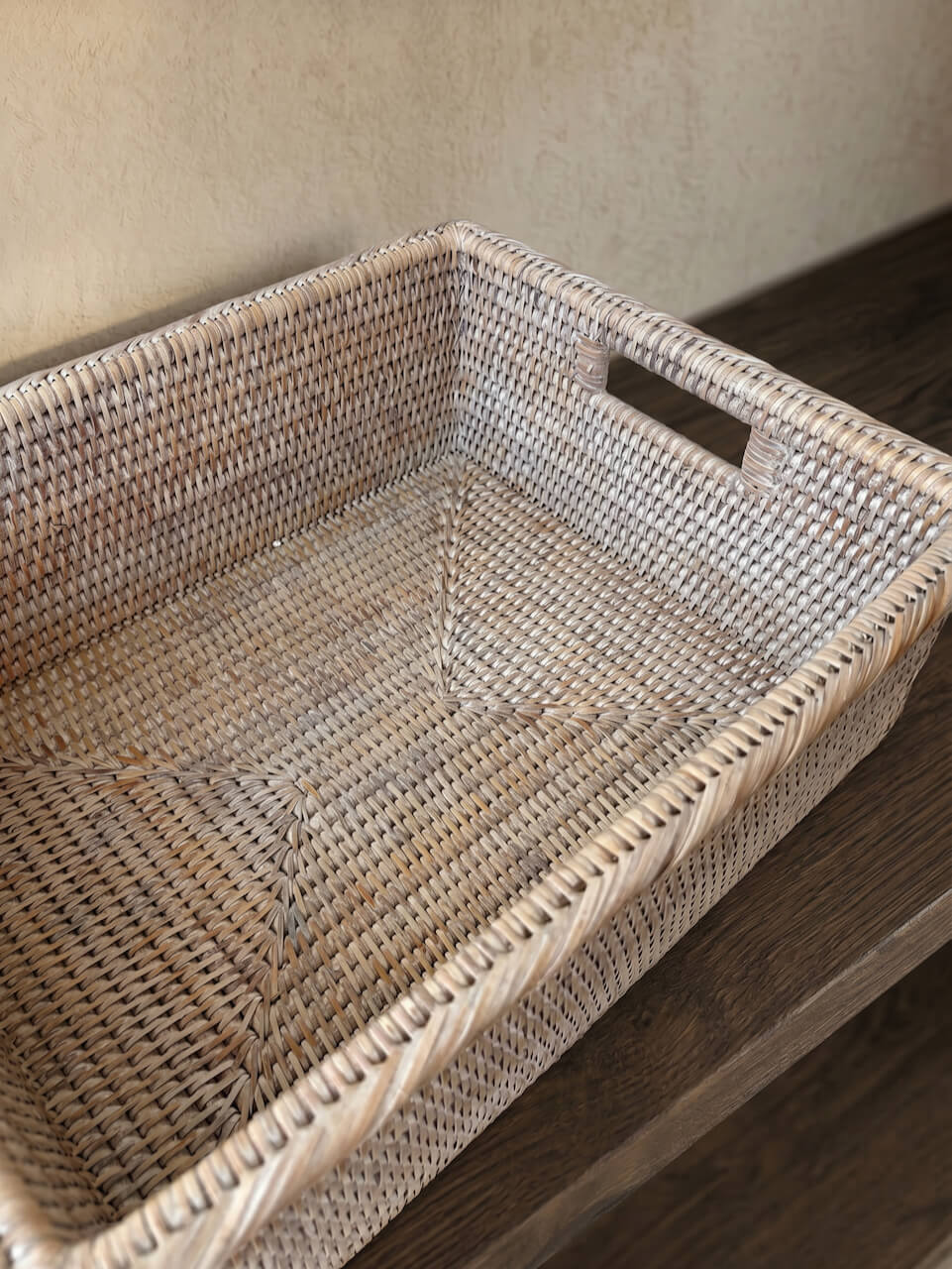 Close-up of a handwoven natural whitewash rattan storage basket with integrated handles, styled on a dark wooden shelf in a warm neutral interior.