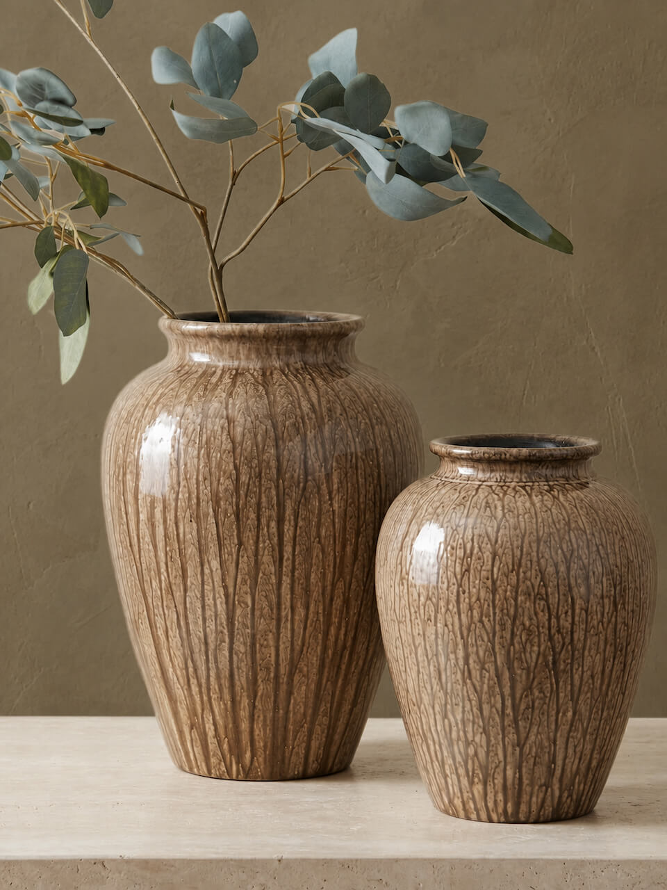Two brown textured ceramic vases with a vertical ribbed pattern, one holding eucalyptus branches, set on a light stone surface against a warm taupe background.
