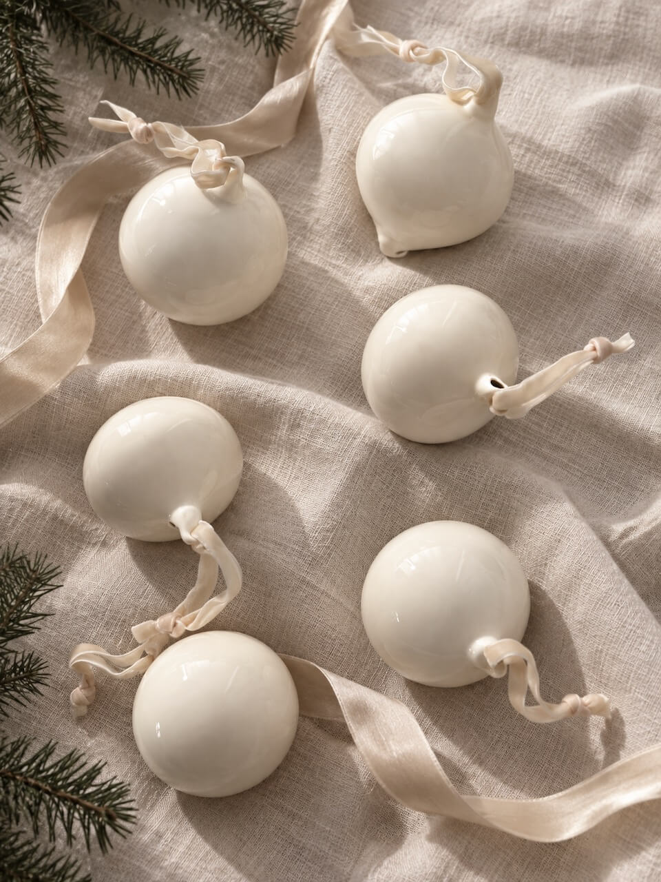 Six glossy white ceramic Christmas ornament pendants with cream ribbons and small wooden tags, arranged on a beige linen fabric with pine branches.