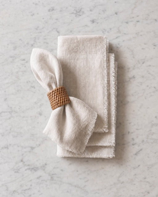 A stack of folded off-white linen napkins with frayed edges, tied with a woven tan napkin ring, placed on a light gray marble surface.