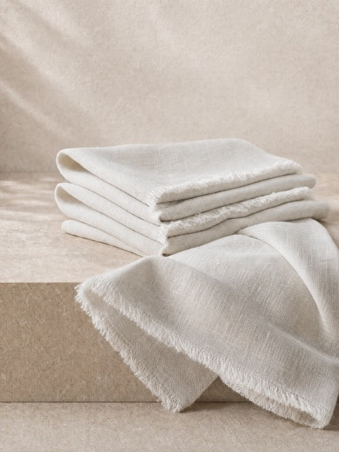 A stack of folded off-white linen napkins with frayed edges, draped over a beige stone block against a matching neutral background.