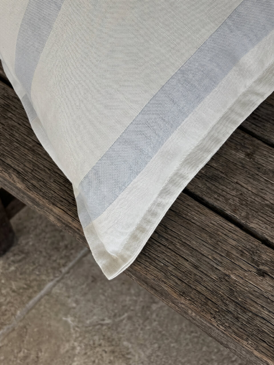 Close-up detail of a luxury blue fog striped cotton linen cushion cover with tailored edging, styled on a rustic timber surface to showcase its soft woven texture and refined craftsmanship.