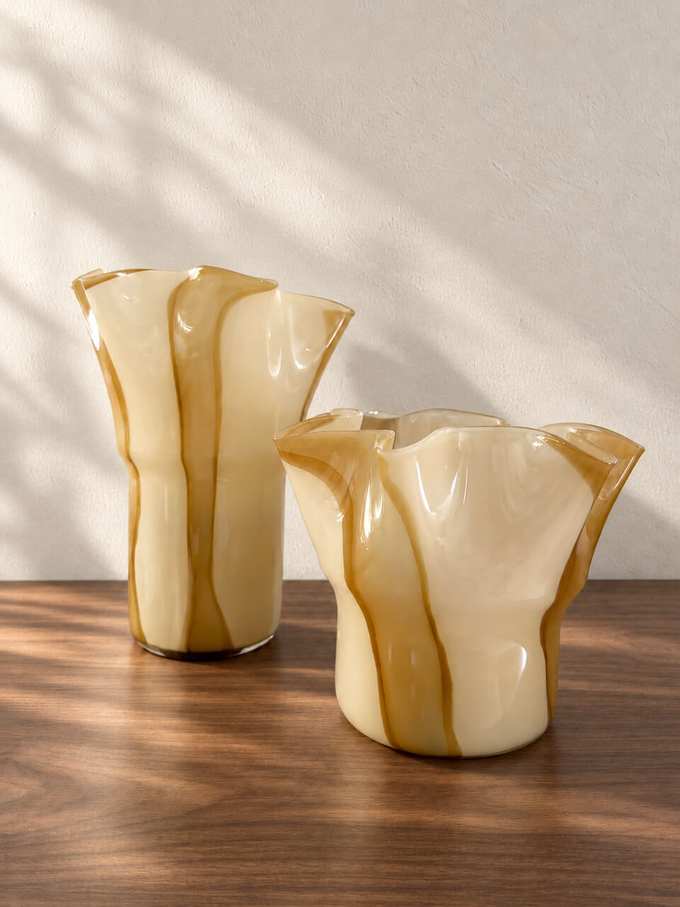 Two fluted glass vases in a warm cream and amber-brown marbled style, displayed on a wooden surface against a textured beige wall with soft window shadow patterns.
