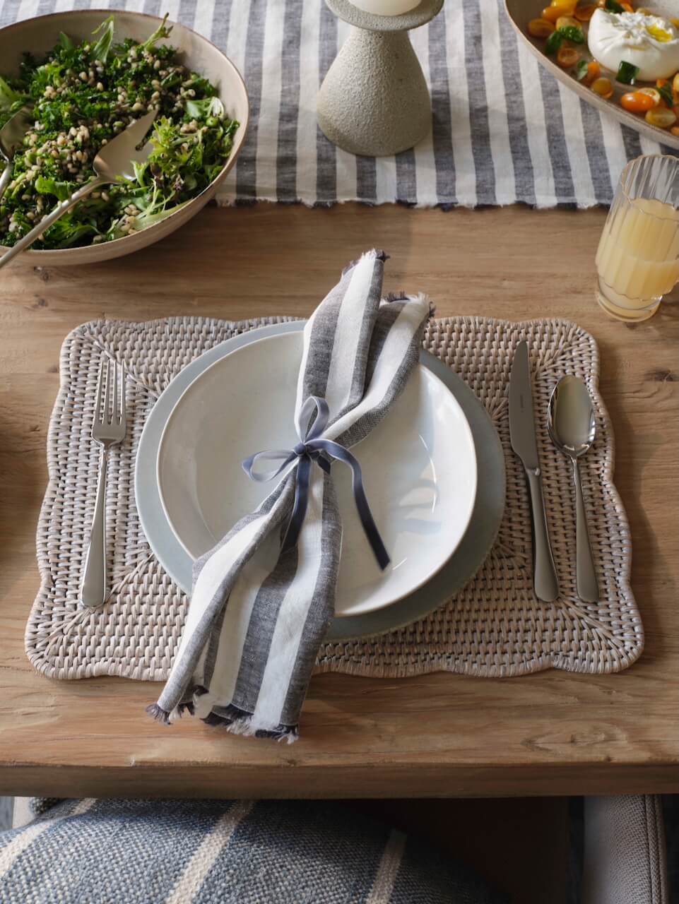 Ceres white rattan placemat with scalloped edge styled on wooden dining table with plates, striped napkin and serving dishes, relaxed table setting UK