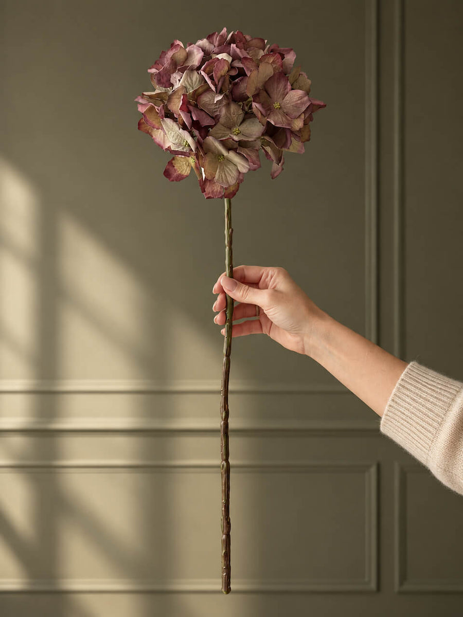 An artificial decorative flower stem with a dusty mauve-pink hydrangea head and a brown twig-like stem, held by a person's hand against a muted olive-green paneled wall.