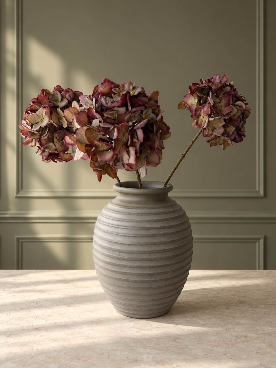 A ribbed ceramic vase in a light warm gray tone, holding artificial hydrangea stems with muted burgundy and tan petals, set on a light stone surface against a paneled sage green wall.