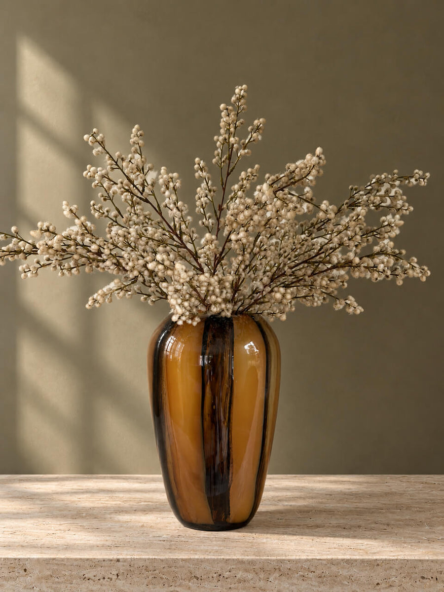 A glossy amber-brown glass vase with dark vertical streaks, filled with dried white baby's breath flowers, set on a light stone surface against a warm taupe wall with soft window shadows.