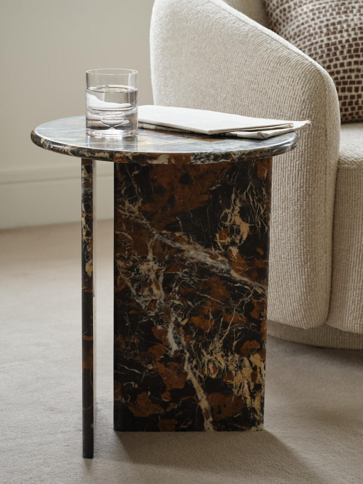 A marble side table with a brown pattern, featuring a glass of water and a remote on top, placed next to a chair in a room with a neutral color scheme.