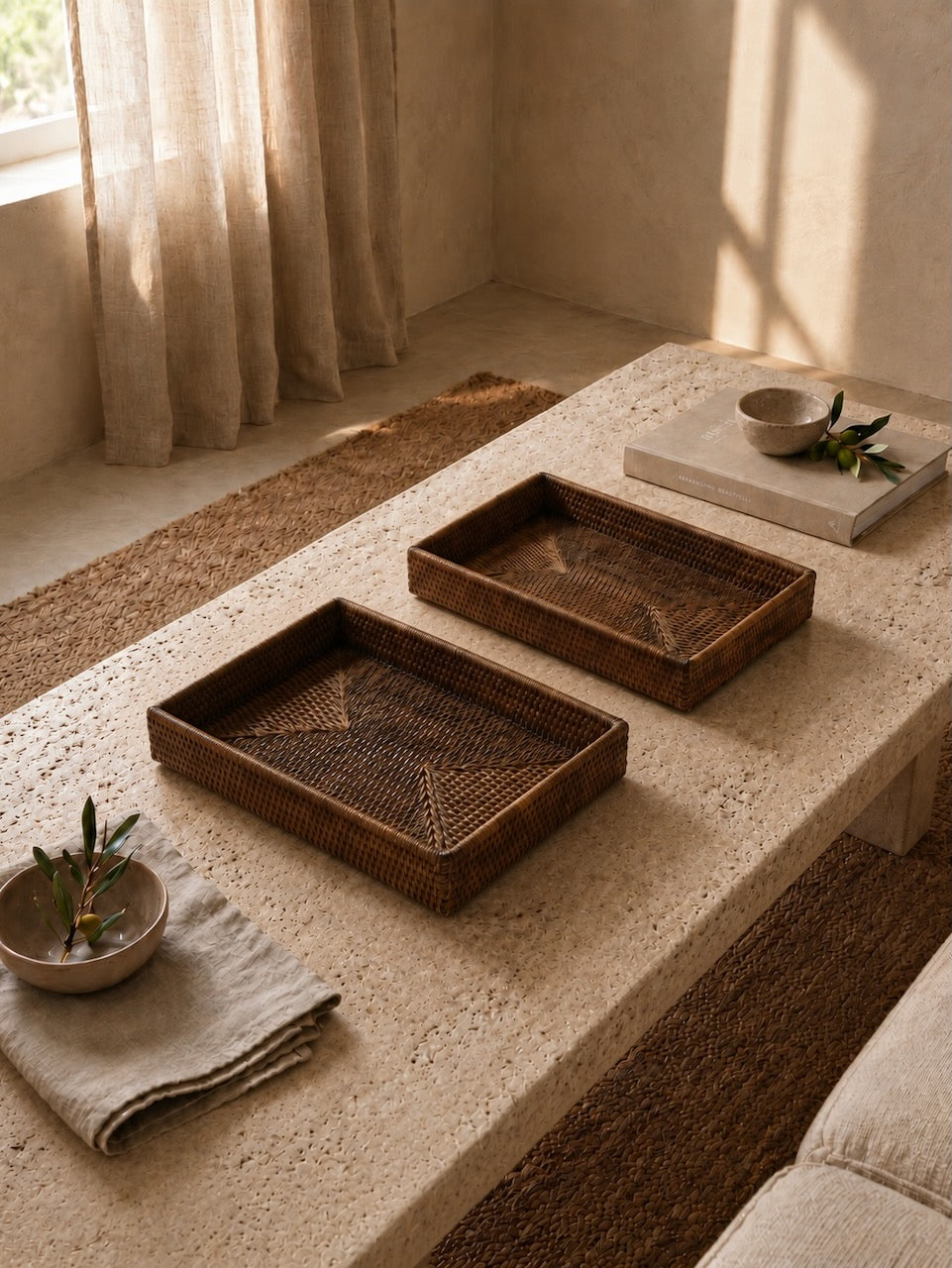 Two small handwoven antique brown rattan trays styled on a textured travertine coffee table in a warm neutral living room with soft linen accents and natural light.