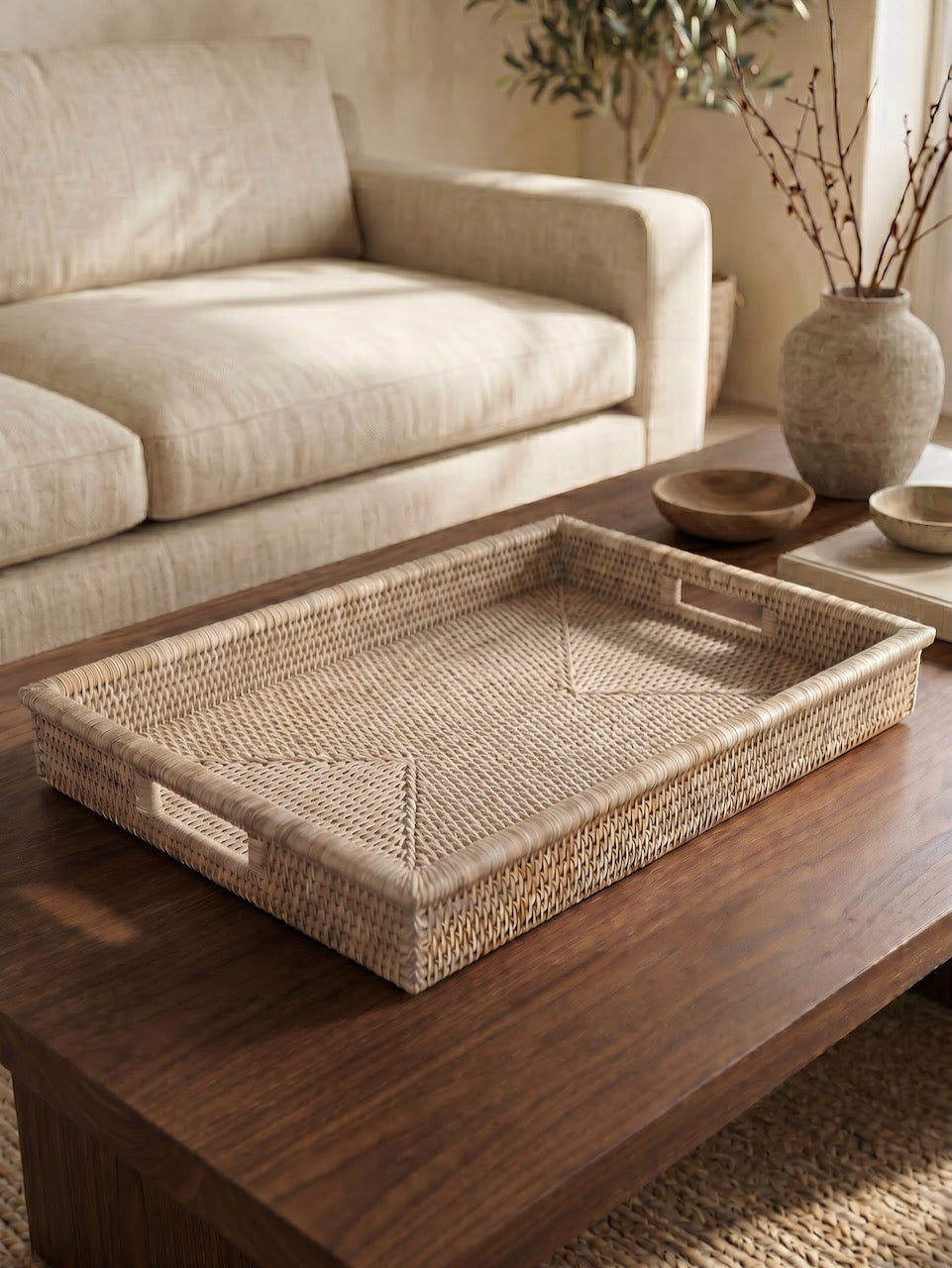 Large rectangular handwoven rattan tray with integrated handles styled on a dark wooden coffee table in a warm, contemporary living room setting.