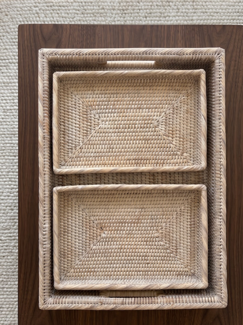 Overhead view of a set of three nesting handwoven rattan trays arranged on a dark walnut coffee table with a textured cream rug background.