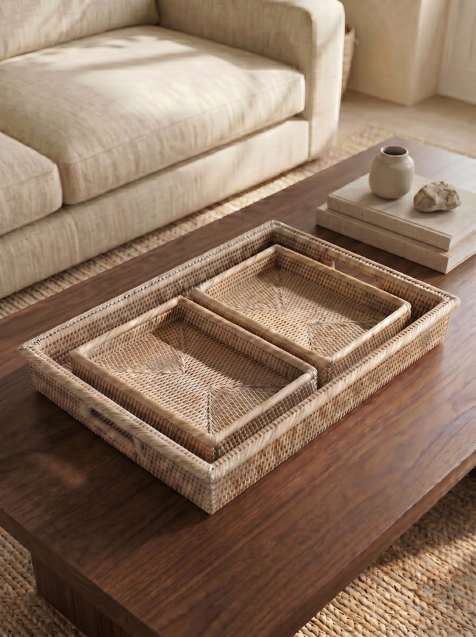 Set of three handwoven natural rattan trays styled on a dark wooden coffee table in a soft neutral living room with linen upholstery and ceramic décor.