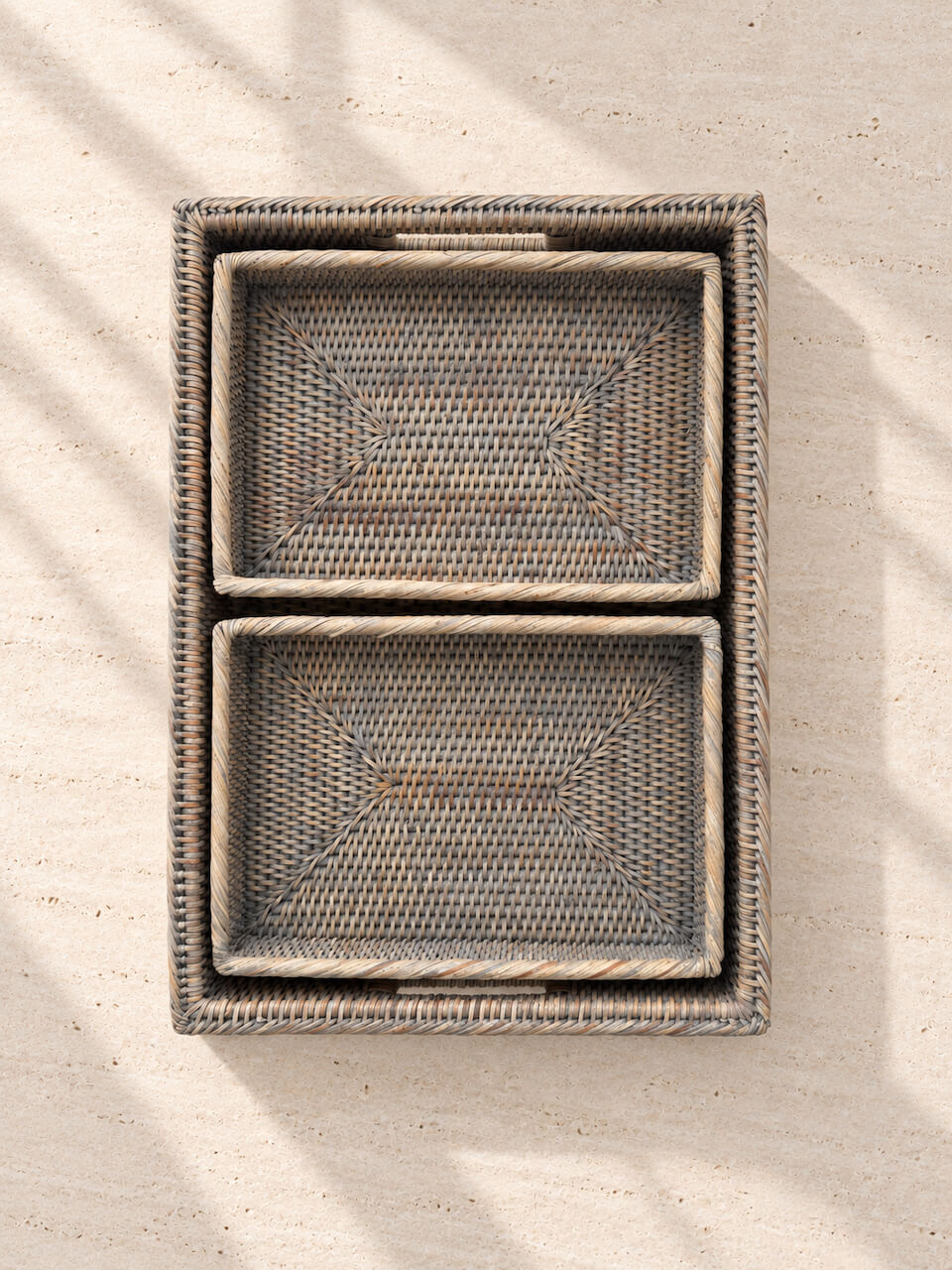 A top-down view of a rectangular woven rattan tray with two nested rectangular trays inside, set against a light beige textured background with soft leaf shadows.