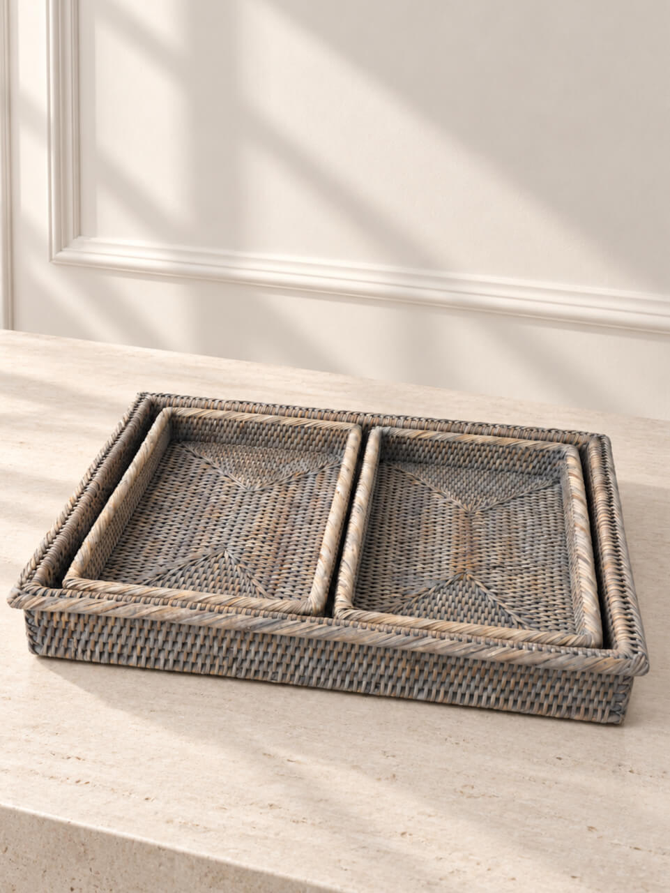 A rectangular woven rattan tray with two nested compartments, made from natural rattan in a light grey tone, resting on a light stone surface against a white paneled wall.