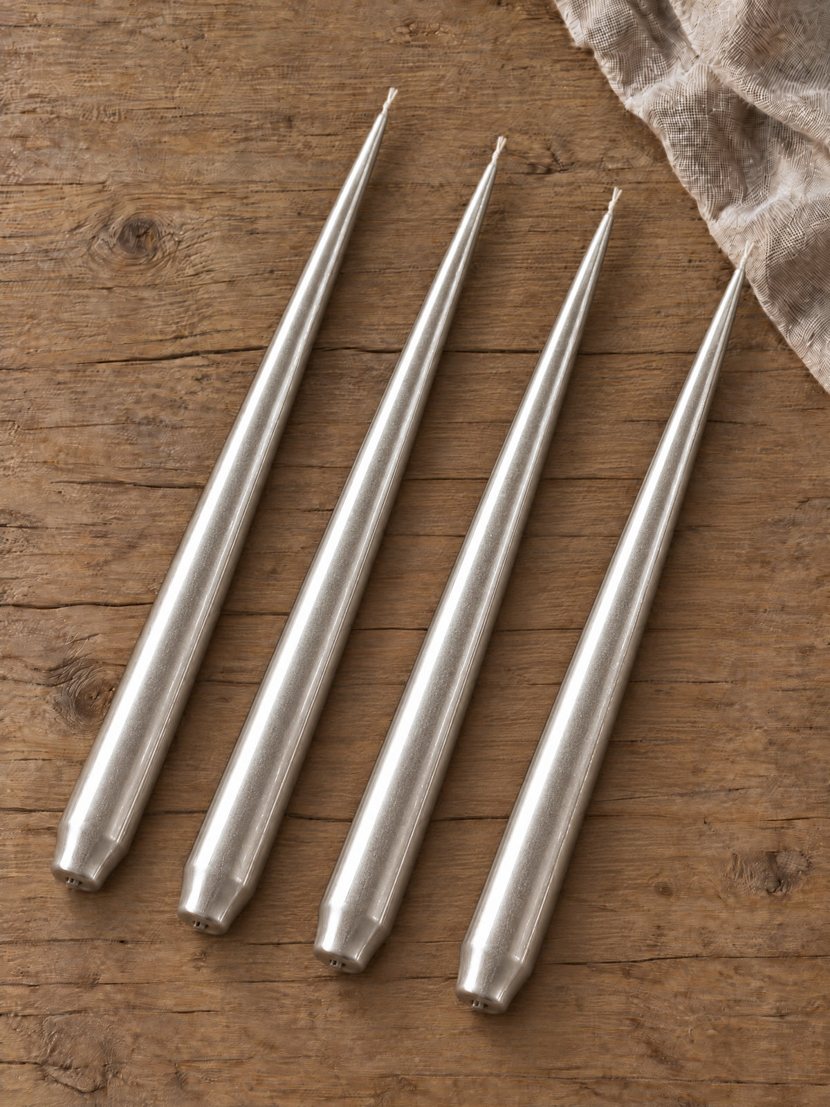 Four unlit metallic silver taper candles arranged diagonally on a wooden table, with folded natural linen at the upper right.