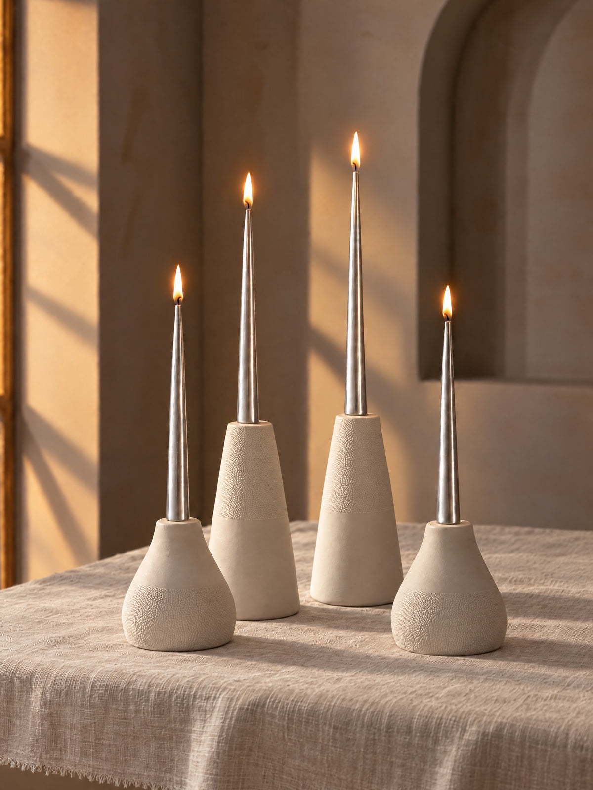 Four lit metallic silver taper candles in ivory textured holders on a putty linen tablecloth, with warm light and an arched wall behind