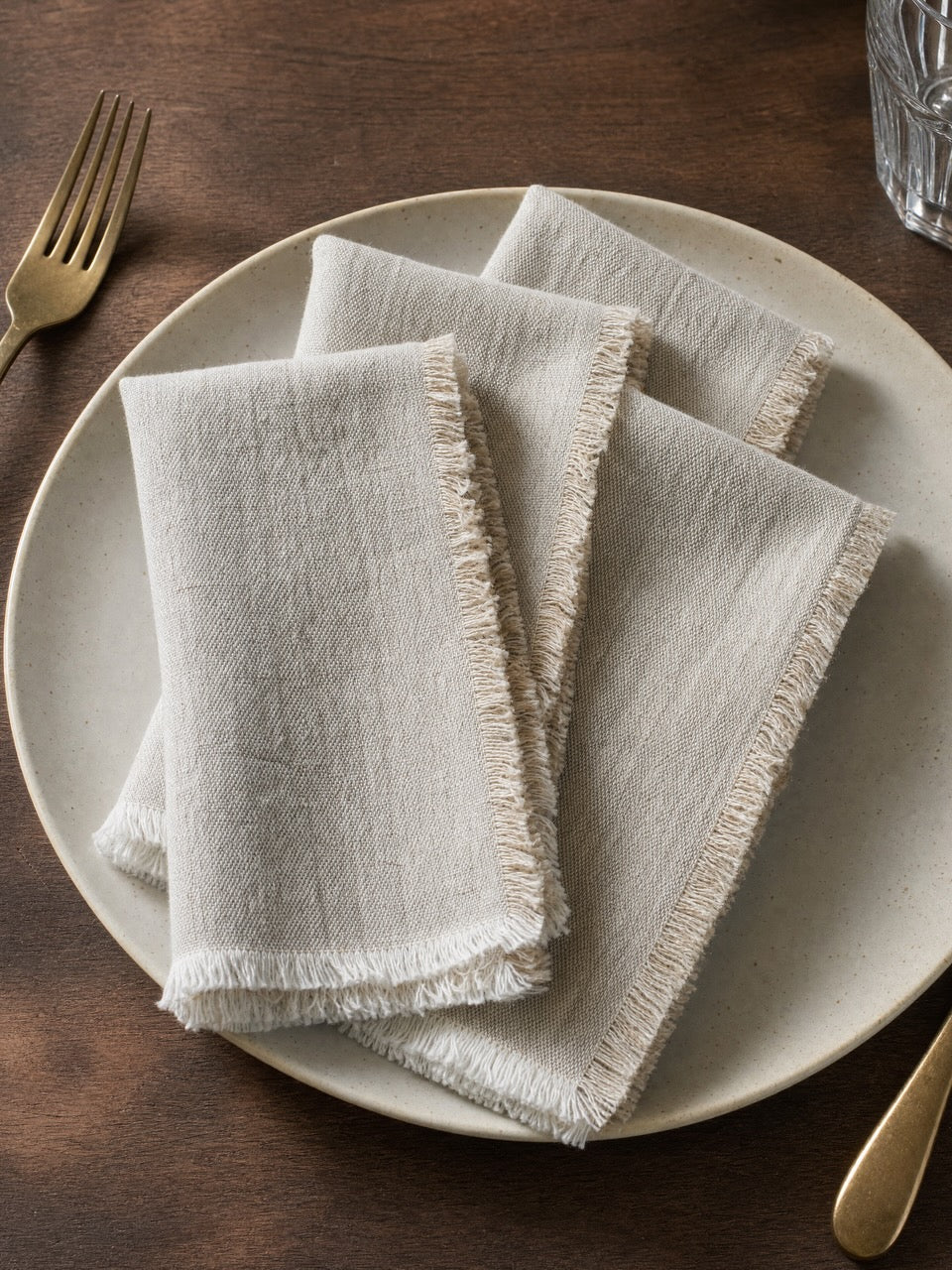 Four folded light putty linen napkins with frayed edges, arranged on a round ceramic plate on a dark wooden table, with a gold fork and glass visible.