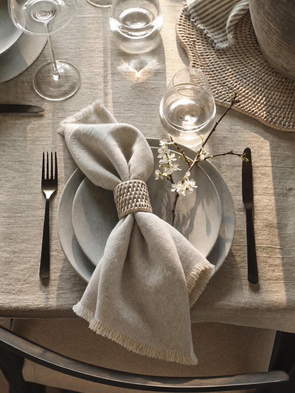 A styled table setting featuring a light Putty linen napkin tied with a woven white rattan napkin ring, layered over Luso plates on a putty linen tablecloth, with silver cutlery and glassware.