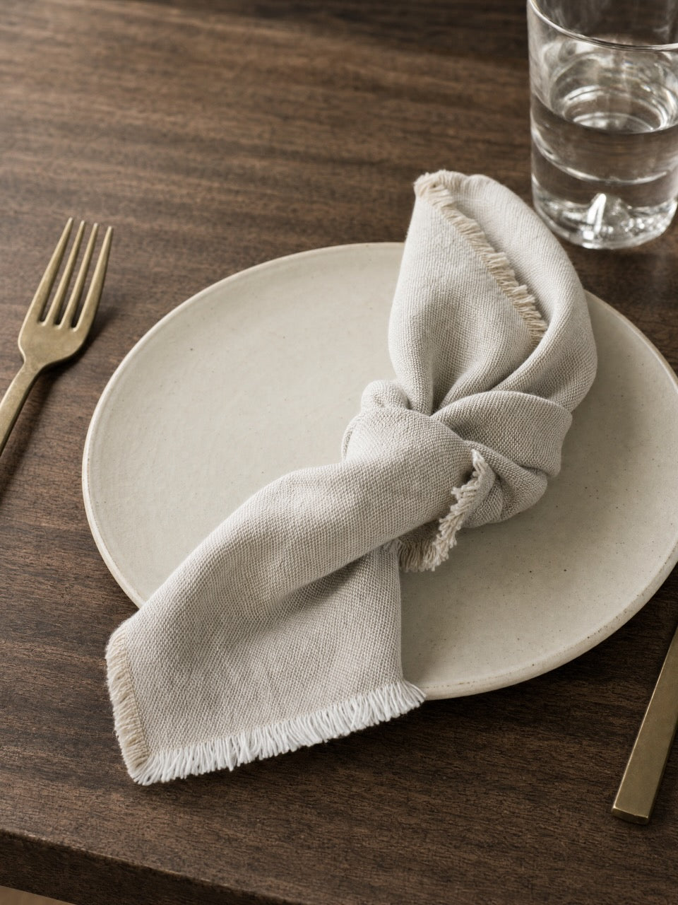 A light putty linen napkin with frayed edges, tied in a knot on a round ceramic plate, set on a dark wood table with a gold fork and a glass of water.