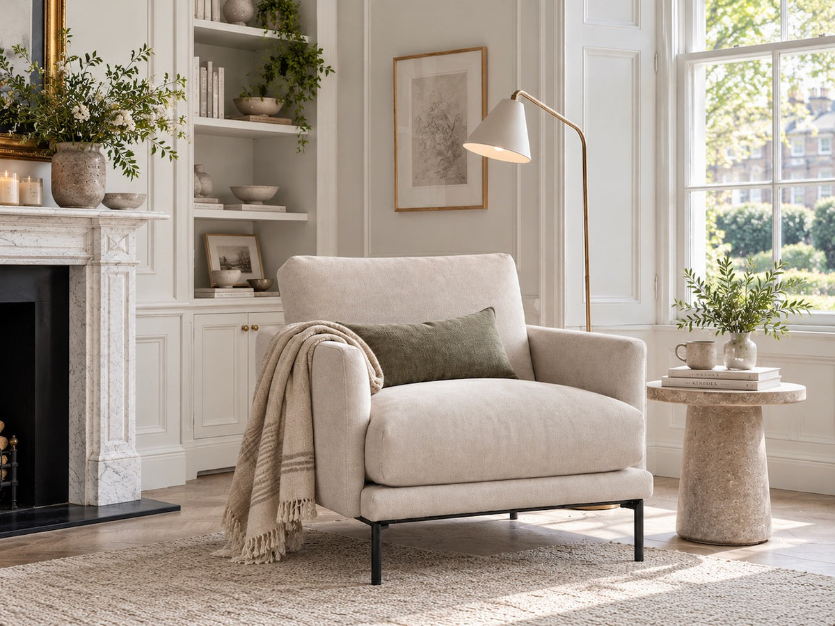 A light beige upholstered armchair with black metal legs in a bright, modern living room. A textured cream throw blanket and a dark green pillow are draped on the chair, which is positioned next to a stone side table, a floor lamp, and a white fireplace with built-in shelves.