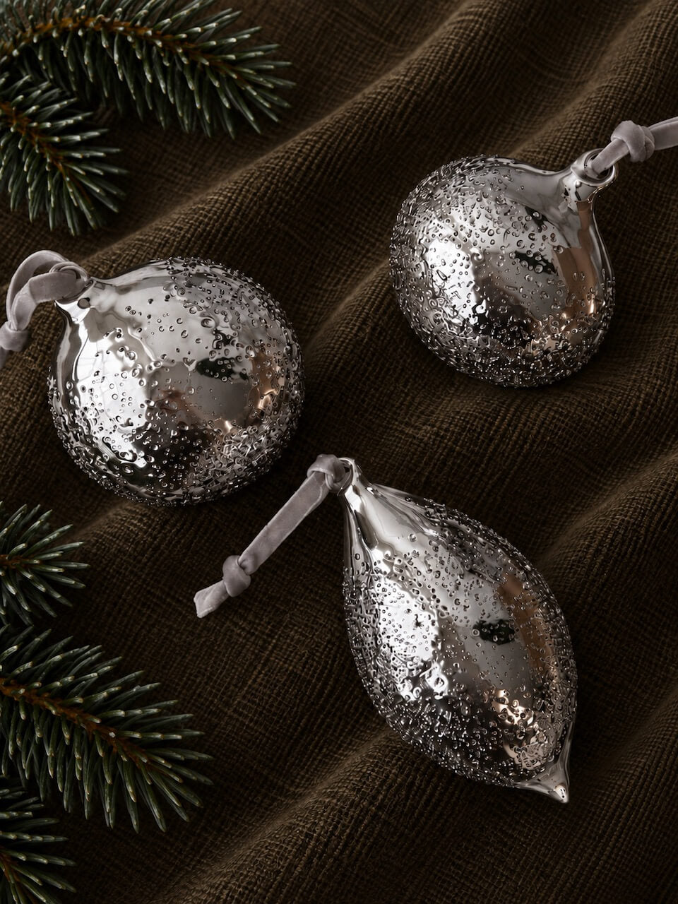 Three silver holiday ornaments with a glittery, snow-dusted finish resting on a dark brown textured fabric background with pine branches.