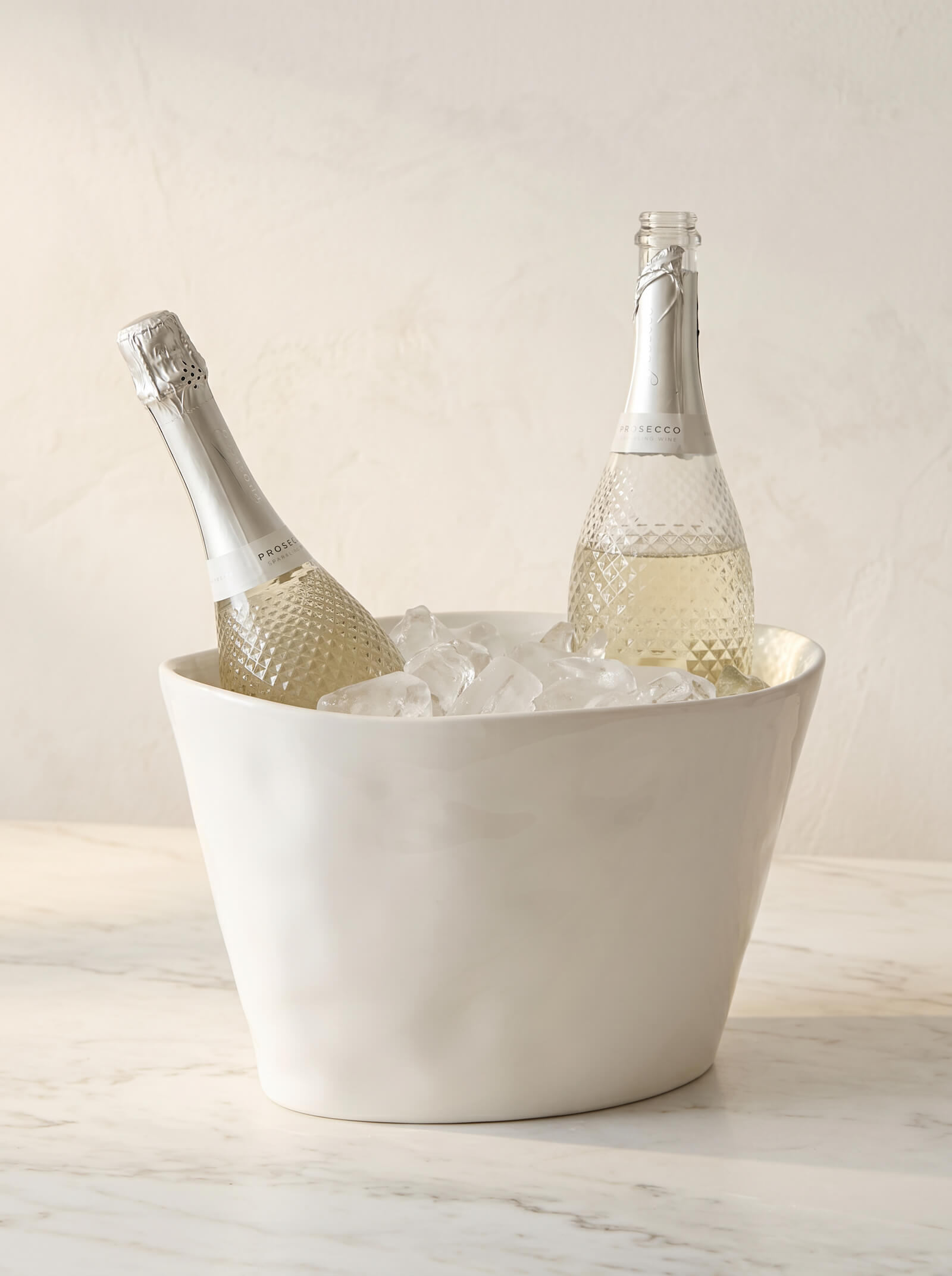 Two bottles of champagne in a white ceramic ice bucket on a marble surface.