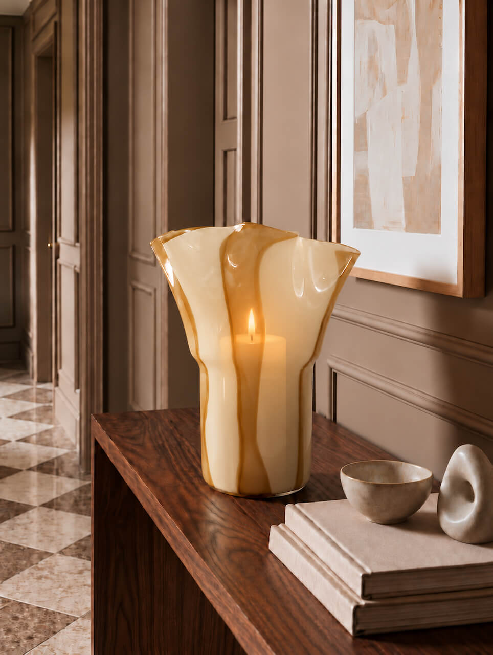 A tall, sculptural flared glass candle holder in warm amber and cream tones, holding a lit candle, displayed on a dark wood console table in an elegant interior hallway.