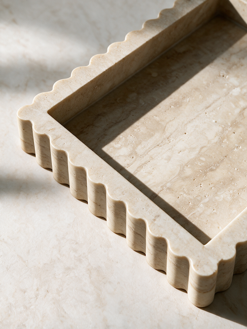 A rectangular stone tray with scalloped edges, shown in a close-up angled view on a matching stone surface. The tray and surface have a warm cream-beige travertine or marble look with natural tonal variation.
